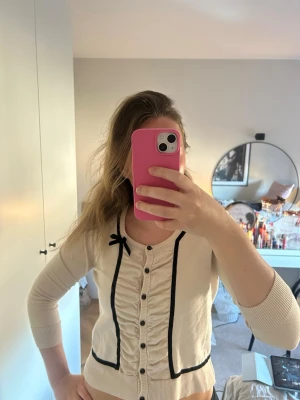 Söt beige kofta med svarta detaljer - Beige kofta med svarta dekorativa kanter och liten rosett vid halsen.