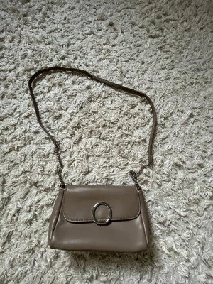 Beige axelväska med silverdetalj - Snygg beige axelväska med kedjerem och stilren silverring på locket. Väskan har en mjuk, slät yta och klassisk rektangulär form. Perfekt för dig som vill ha en enkel men trendig accessoar till din outfit.