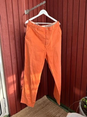 Orange chinos Polo från Ralph Lauren - Snygga orange chinos från Ralph Lauren med raka ben och klassisk passform. Byxorna har två sidofickor fram och två bakfickor med knapp. Tillverkade i ett lätt och bekvämt bomullsmaterial som passar perfekt för en avslappnad stil.