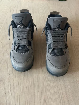 Gråa Air Jordan 4 Retro sneakers - Säljer ett par gråa Air Jordan 4 Retro sneakers med svarta och mörkgrå detaljer. Skorna har klassisk mesh på sidorna, mockaliknande material och ikonisk Jumpman-logga på plösen. Perfekta för dig som gillar streetwear och snygga detaljer.