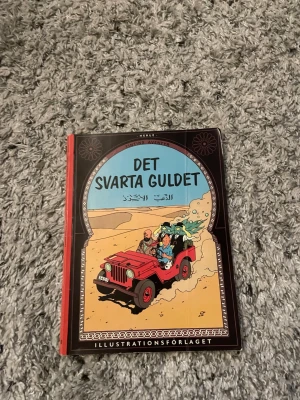 Tintin: Det svarta guldet - Säljer seriealbumet 'Det svarta guldet' av Hergé, en klassisk Tintin-bok med färgglada illustrationer och hårda pärmar. Omslaget är svart, blått och rött med motiv av Tintin och hans vänner i en röd jeep i öknen. Perfekt för dig som gillar äventyr och serier.