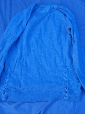 Grå sweatshirt från Ellos - Grå sweatshirt från Ellos med långa ärmar och rund hals. Tröjan har dekorativa snörningsdetaljer längs sidorna och ribbade muddar vid nederkant. Perfekt för en avslappnad och trendig look.