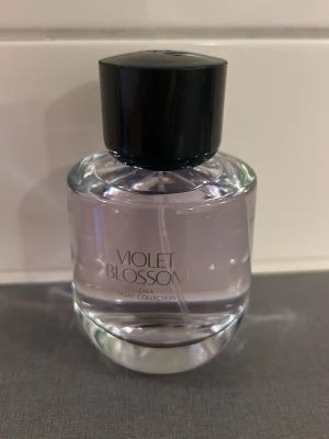 Zara Violet Blossom parfym - Zara Violet Blossom parfym från Night Collection med en stilren, rund glasflaska och svart lock. Doften har en lila ton och flaskan har en modern design med enkel text. Perfekt för dig som gillar eleganta och fräscha parfymer.