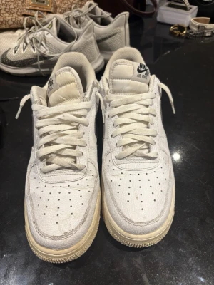 Nike Air Force 1 vita sneakers - Klassiska Nike Air Force 1 sneakers i vitt läder med svart swoosh på sidan. Skorna har perforerad tå, vit sula och snörning framtill. Den ikoniska modellen har en chunky siluett och passar perfekt till streetwear-stilen.