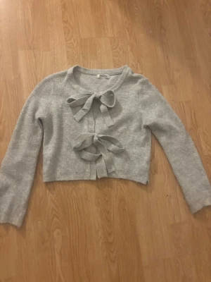 Ljusgrå stickad kofta med rosetter - Superfin ljusgrå stickad kofta med två stora rosetter framtill. Perfekt för att ge din outfit en söt och trendig touch.