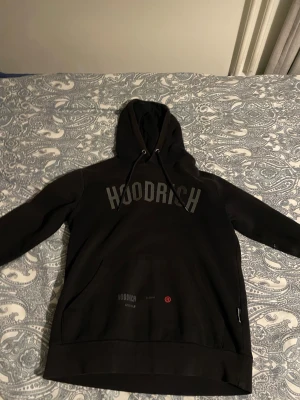 Svart hoodie från Hoodrich - Cool svart hoodie från Hoodrich med stor logga på bröstet och tryck på ryggen. Klassisk huva med dragsnören och en stor magficka. Materialet är mjukt och bekvämt, perfekt för en avslappnad streetwear-look. Snygga detaljer och modern passform.