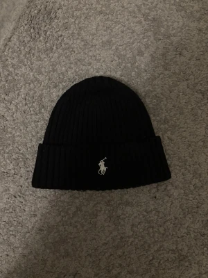 Svart ribbad mössa från Polo Ralph Lauren - Svart ribbstickad mössa från Polo Ralph Lauren med klassisk uppvikt kant och broderad vit logotyp framtill. Perfekt för att hålla värmen och ge en stilren look. Materialet är mjukt och stretchigt, vilket gör den bekväm att bära.