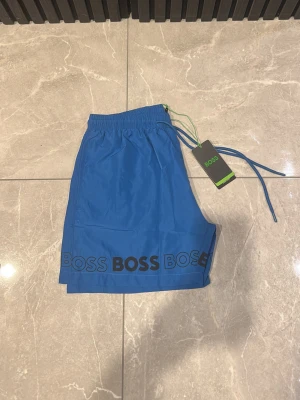 Blå badbyxor från BOSS - Snygga blå badbyxor från BOSS med elastisk midja och dragsko. BOSS-loggan är tryckt flera gånger längs nederkanten i svart. Materialet är lätt och snabbtorkande, perfekt för strand eller pool.