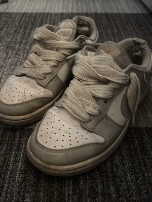 Nike Dunk Low vita/grå sneakers - Nike Dunk Low sneakers i vitt och ljusgrått med klassisk swoosh på sidan. Skorna har perforerad tåbox, tjocka vita skosnören och vadderad plös. Tillverkade i läder och textil för en clean och tidlös look.