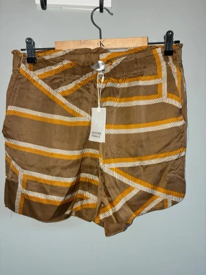 Bruna mönstrade shorts Second Female - Säljer ett par bruna shorts från Second Female med coolt grafiskt mönster i vitt och orange. Shortsen har elastisk midja och är tillverkade i ett mjukt, glansigt material som känns lyxigt mot huden. Perfekta för varma dagar och ger en trendig touch till din outfit.