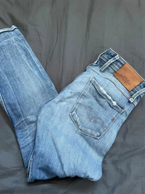 Blå slitna jeans från Replay - Säljer ett par blå jeans från Replay med slitna detaljer och snygga slitningar på både fram- och baksida. Klassisk femficksmodell med normal passform och dragkedja. Perfekta för en avslappnad och trendig look.