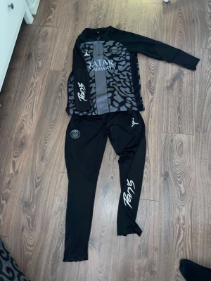 PSG träningsset Jordan x Paris - Snyggt svart träningsset från PSG och Jordan med gråa detaljer och mönster på tröjan. Setet består av långärmad tröja och matchande byxor med Paris-logga och Jordan-logga. Perfekt för fotbollsträning eller chill. Materialet är troligtvis polyester för bästa komfort. Jag är 165 och den passar perfekt!