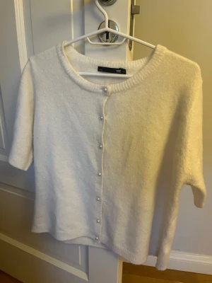 Vit kofta med pärlknappar - Söt vit kofta från Lager157 med korta ärmar och rund halsringning. Koftan har dekorativa pärlknappar framtill och är stickad i ett mjukt material som känns fluffigt. Perfekt för att lyfta en enkel outfit med en elegant touch.