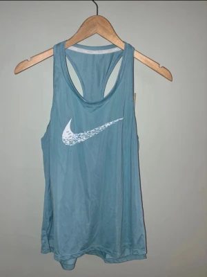 Blått träningslinne från Nike - Snyggt blått träningslinne från Nike med vitt swoosh-tryck på bröstet. Linnet har brottarrygg och är gjort i ett lätt, snabbtorkande material som passar perfekt till gymmet eller löpning.