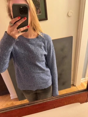 Blå stickad tröja med rund hals - Mysig blåmelerad stickad tröja med rund halsringning och långa ärmar. Tröjan har en enkel, klassisk design och ribbstickade muddar vid ärmslut och nederkant. Perfekt för lager på lager och passar till både jeans och kjol. 