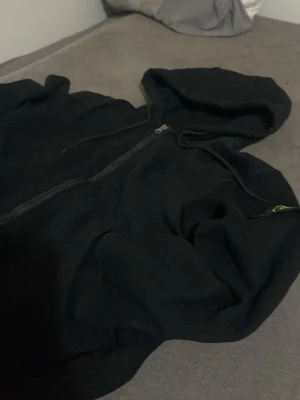 Svart hoodie från Stone Island - Säljer en svart hoodie från Stone Island med dragkedja framtill och klassisk huva. Ärmen har den ikoniska Stone Island-loggan i gult och grönt. Hoodien är i mjukt material och har en clean, stilren look som passar perfekt till streetwear.