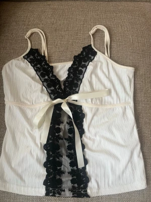 Vit linnetopp med spets och rosett - Supersöt vit linnetopp med smala axelband, svart spetsdetalj framtill och en dekorativ rosett i midjan. Toppen har en lätt ribbad struktur och är perfekt för dig som gillar romantiska detaljer.🤍Storlek M-L