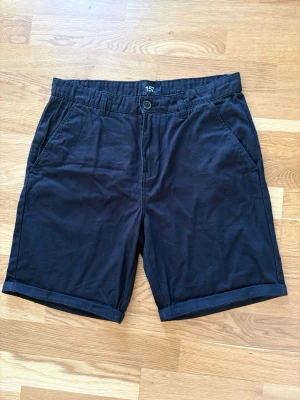 Svarta bomullsshorts i chinosmodell - Stilrena shorts. Har två sidofickor fram, två bakfickor med knapp och hällor för bälte. Perfekta för varma dagar och enkel att matcha med olika outfits. Använda en gång. Uppviket behöver man inte ha om man hellre vill ha raka. Det är inte fastsytt.  Uppviket är 2 cm. Benlängd med uppviket 48 cm, annars 50 cm. Innerbenslängd med uppvik 21 cm, annars 23. Använda två gånger.