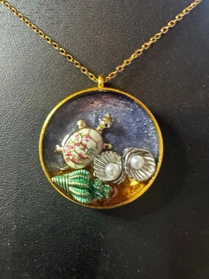 Halsband med sköldpadda och pärldetaljer - Unikt halsband med runt hängsmycke i guldton. Smycket har en söt sköldpadda med blommönster, två snäckor med pärlor och en grön snäcka. Kedjan är tunn och guldfärgad. Perfekt för dig som gillar havsinspirerade accessoarer och detaljer.