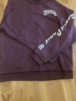 Vinröd sweatshirt från Hollister - Vinröd sweatshirt från Hollister med tryckt vitt Hollister California-märke på bröstet och text längs ena ärmen. Tröjan har rund halsringning och ribbade muddar vid ärmslut och nederkant. Perfekt för en avslappnad och trendig look.