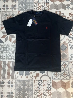 Svart t-shirt från Polo Ralph Lauren - Svart t-shirt från Polo Ralph Lauren med klassisk passform och röd broderad logga på bröstet. Tillverkad i mjuk bomull och har rund halsringning samt korta ärmar. Perfekt för en clean och stilren look.