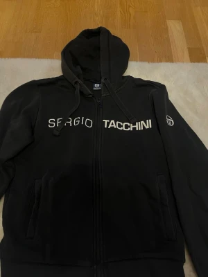 Svart hoodie från Sergio Tacchini - Svart hoodie med dragkedja från Sergio Tacchini. Stor vit logga och text framtill samt logotyp på ärmen. Utrustad med huva och dragsnören. Mjuk och bekväm, perfekt för en avslappnad stil.
