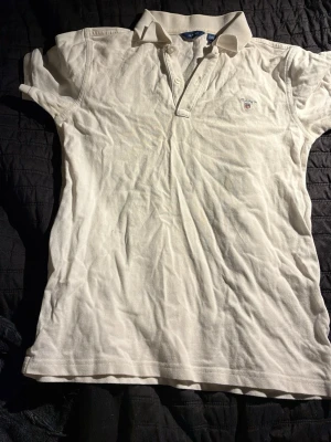 Vit pikétröja från Gant - Klassisk vit pikétröja från Gant med korta ärmar och krage. Tröjan har knappar framtill och en liten broderad logga på bröstet. Perfekt för en stilren och sportig look.