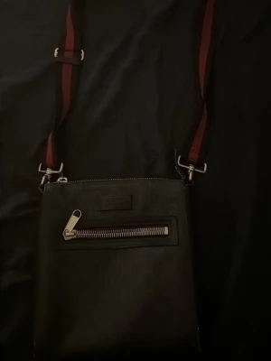 Svart axelväska från Gucci - Snygg svart axelväska från Gucci med justerbar axelrem i rött och svart. Väskan har en framficka med dragkedja och diskret logga på framsidan. Tillverkad i skinn med detaljer i metall och tygfoder invändigt. Perfekt för att bära det viktigaste med stil.äkta!