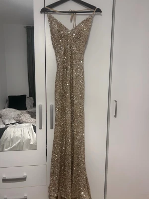 Glittrig guldig långklänning - Säljer en glittrig guldig långklänning med smala axelband och v-ringning. Klänningen har en slits framtill och är täckt av paljetter som ger ett riktigt lyxigt intryck. Perfekt för dig som vill sticka ut på bal eller fest! Materialet känns mjukt och följsamt. Öppning i rygg och knytband i nacken där du kan välja storlek. (Jag är 155cm lång) finns bilder 