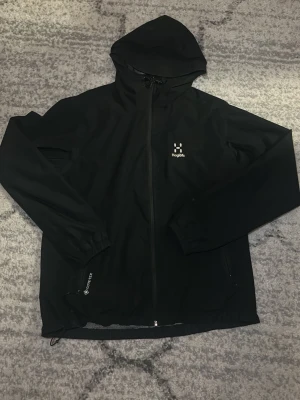 Svart vindjacka från Haglöfs - Snygg svart vindjacka från Haglöfs med huva och hel dragkedja framtill. Jackan har två sidofickor med dragkedja, justerbar nederkant och elastiska muddar vid ärmsluten. Tillverkad i ett lätt och vindtätt material, perfekt för blåsiga dagar.