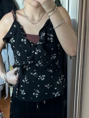 Svart blommig linnetopp med volang - Supersöt svart linnetopp med små blommor i vitt och rosa. Toppen har tunna axelband och volangdetalj framtill som ger en romantisk vibe. Perfekt för varma dagar eller lager på lager. Materialet känns lätt och luftigt.