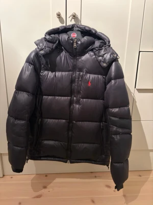 Svart dunjacka från Polo Ralph Lauren - Snygg svart dunjacka från Polo Ralph Lauren med avtagbar huva och dragkedja framtill. Jackan har två sidofickor med dragkedja och den klassiska röda loggan broderad på bröstet. Perfekt för kalla vinterdagar och riktigt skön att bära.storlek L/M passar för båda storlekarna!
