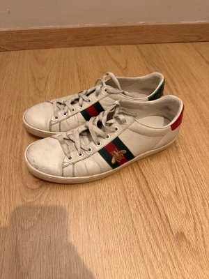 Gucci Ace vita sneakers med bi - Gucci Ace sneakers i vitt läder med klassiska gröna och röda ränder på sidan och broderad guldfärgad bi. Låg modell med vita snören och röd samt grön detalj på hälen. Perfekta för dig som gillar lyxiga och trendiga sneakers.