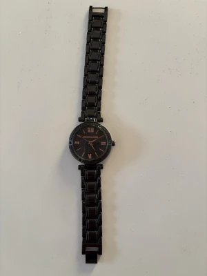 Svart klocka från Michael Kors - Stilren svart klocka från Michael Kors med metallänk och urtavla i svart med romerska siffror och detaljer i guld. Klassisk rund form och elegant design som passar till många olika looks. Behöver nytt batteri