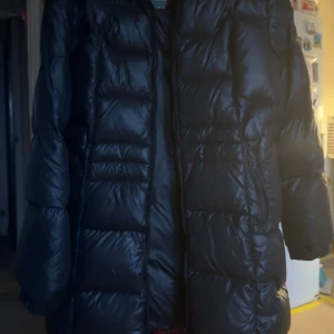 Svart lång dunjacka från 8848 Altitude - Säljer en svart, lång dunjacka från 8848 Altitude med snygg quiltad design och dragkedja framtill. Jackan har lång ärm och är perfekt för kalla dagar. Märket är broderat på ärmen och materialet är glansigt.  Storleken är 44 men kan ej skriva det på annonsen. Knappt använd 