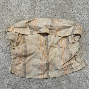 Randig tubtopp från Divided H&M - Säljer en beige och ljusbrun randig tubtopp från Divided H&M. Toppen är axelbandslös med en bred volang upptill och har dragkedja i sidan. Perfekt för dig som gillar en chill och trendig stil.