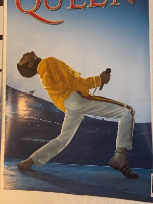 Queen affisch med ikoniskt motiv - Säljer en affisch med ett ikoniskt motiv av Queen. Affischen har en färgstark bild med blå bakgrund och en person i gul jacka och vita byxor som håller en mikrofon. Perfekt för musikälskare och fans av klassisk rock.