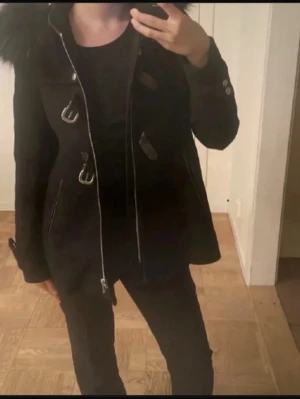 Svart duffelkappa med fuskpäls - Stilren svart duffelkappa med dragkedja och dekorativa spännen framtill. Kappan har stor huva med fuskpäls och knappar vid ärmsluten. Perfekt för kyliga dagar och ger en snygg siluett. Passar till både jeans och klänning.