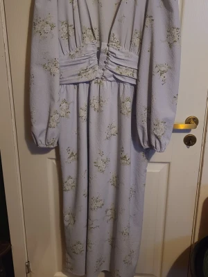 Ljuslila blommig långklänning - Superfin långklänning i ljuslila med blommigt mönster och djup v-ringning. Klänningen har långa puffärmar och markerad midja med veckade detaljer. Perfekt för dig som gillar romantisk och trendig stil.