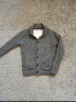 Grå kabelstickad cardigsn - Mysig grå kofta med kabelstickat mönster. Koftan har knäppning med svarta knappar framtill, ribbade muddar och två fickor. Perfekt för lager på lager och en stilren look.