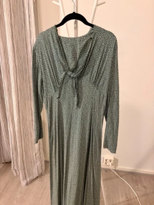 Grön långklänning med knyt i nacken - Snygg grön långklänning med småprickigt mönster och knyt i nacken. Klänningen har trekvartsärm och ett mjukt, följsamt tyg som ger en avslappnad men ändå elegant look.