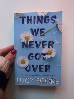 Things We Never Got Over - En skönlitterär bok sopad i ljusblått omslag med prästkragar och mörkblå text. Boken är skriven av Lucy Score och har en modern, ungdomlig design. Perfekt för dig som gillar romantik och drama i småstadsmiljö.
