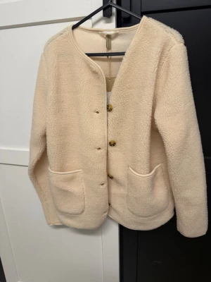 Beige teddyjacka med stora fickor - Mysig beige teddyjacka med rund halsringning och knäppning framtill. 
