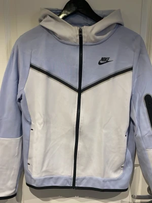 Nike Tech Fleece hoodie ljusblå/vit - Nike Tech Fleece hoodie i ljusblå och vitt med svarta detaljer och dragkedja framtill. Snygg design med svart Nike-logga på bröstet och praktisk ficka med dragkedja på ärmen. Perfekt för en sportig och trendig look.