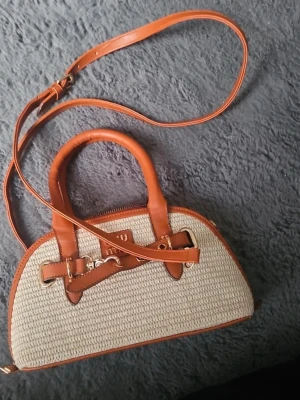Beige och brun handväska i rotting - Snygg handväska i beige flätad rotting med bruna läderdetaljer och guldfärgade metalldelar. Väskan har både handtag och justerbar axelrem, samt dragkedja upptill. Perfekt accessoar för en trendig look.
