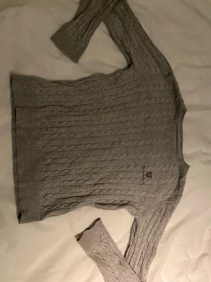 Grå kabelstickad tröja Bondelid - Snygg grå kabelstickad tröja från Bondelid med broderad logga på bröstet. Tröjan har rund halsringning, långa ärmar och ribbade muddar vid ärmslut och nederkant. Perfekt för dig som gillar klassisk och stilren look.