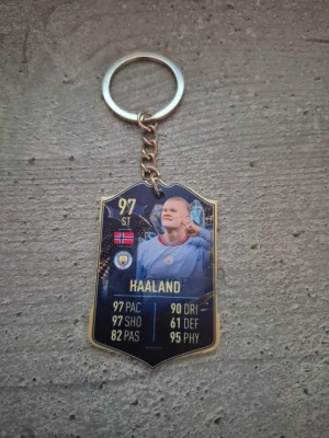 Nyckelring med Haaland-motiv - Cool nyckelring med motiv av fotbollsspelaren Haaland, inspirerad av FIFA Ultimate Team-kort. Designen har blå och guld färger, statistik och klubbmärke. Perfekt för fotbollsälskare som vill ha något unikt till sina nycklar.