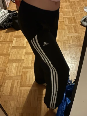 Svarta Adidassweatpants med vita ränder - Säljer ett par svarta mjukisbyxor från Adidas med klassiska vita ränder längs benen och Adidas-logga på låret. Byxorna har en avslappnad passform och resår i midjan. Perfekta för chill dagar eller träning.