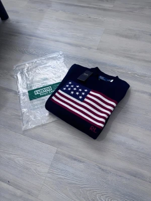Mörkblå stickad tröja Polo Ralph Lauren - Mörkblå stickad tröja från Polo Ralph Lauren med amerikansk flagga i rött, vitt och blått på bröstet. Rund halsringning och RL-broderi i rött nedtill. Klassisk och stilren design, perfekt för dig som gillar preppy mode.