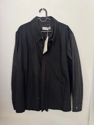 Svart rutig dubbel skjorta från Our Legacy - Säljer en svart skjorta från Our Legacy med rutiga detaljer på ärmar och insida. Skjortan har klassisk krage, knappar framtill och är tillverkad i tokgosig bomull. Perfekt för dig som gillar en clean men ändå unik stil. Den är double layered.